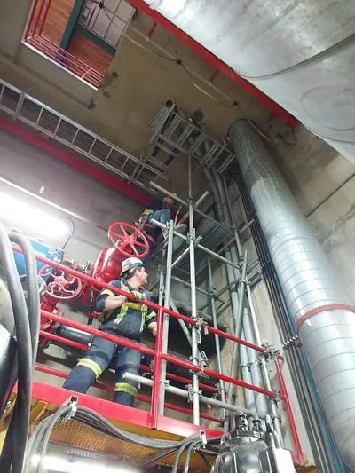 A man in yellow jacket on scaffolding near pipes.